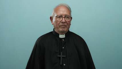 Senior priest in black robe ponders solemnly against a blue isolated background wall, capturing a thoughtful expression of introspection and wisdom.