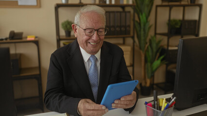 Senior businessman in a suit having a video call on a tablet, smiling in a modern office setting with plants and office supplies creating a professional but warm atmosphere.