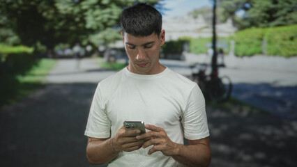 Fototapeta premium Man in white tshirt holding smartphone with hands texting in sunny green park; serene concentration.