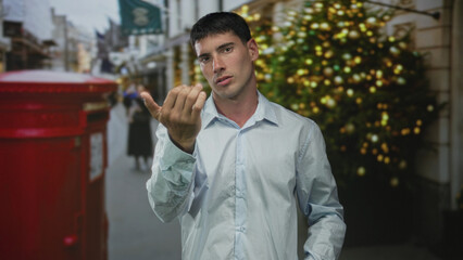 Young hispanic man wearing light blue shirt looking directly and beckoning hand on street;...