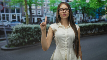 Teenage woman in white sleeveless top and glasses showing two fingers peace sign while counting on a tree lined urban street; playful.