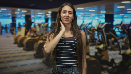 Teen girl wearing striped tank top and braces holding her neck with grimace beside treadmills in gym; discomfort.