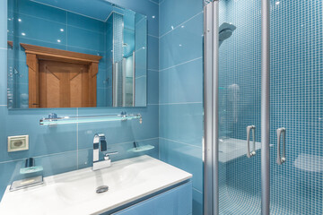 A blue tiled bathroom features a sink with a mirror above, a glass shower, and wooden accents. The...