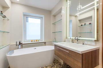 A bathroom features a freestanding tub beside a large window. There are shelves for storage and a sink with a mirror. The space is bright and organized, with a clean design