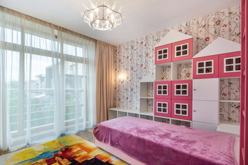 Fototapeta premium This room features a large bed covered with a purple blanket. A pink shelf with house shapes stands against the wall, and big windows let in light