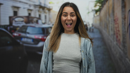Fototapeta premium Young hispanic woman with hands on cheeks smiling on a city street between parked cars; surprise energy.