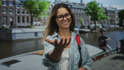 Woman holds out hand on canal street in amsterdam with boat and buildings visible; invitation travel welcoming.