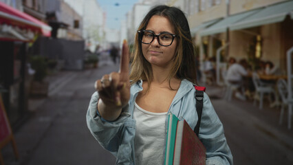 Young hispanic woman pointing index finger toward camera on a street while holding notebooks and a backpack, standing by a restaurant terrace; disapproval.