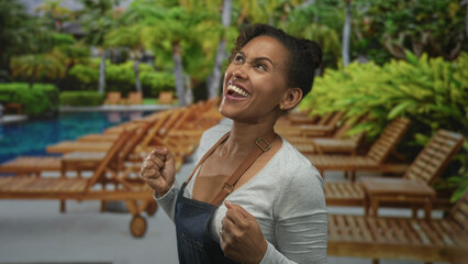 Woman cheering with raised fists and wide smile by a hotel pool building under palm trees; celebration joy.