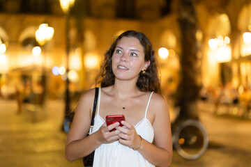 Young woman with a mobile phone in her hands near night bars and restaurants, she answers messages to friends and calls a taxi on the app.