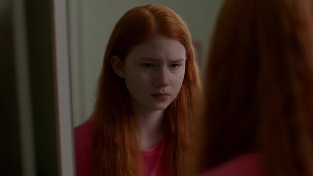 A redhead girl looking in a mirror with a sad facial expression. Reflection of the girl. Emotional distress shown Stock Video