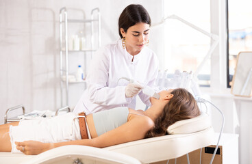 Focused young female cosmetologist performing facial skin cleansing with ultrasonic shovel on woman to enhance efficacy of skincare products and improving skin texture. Modern hardware cosmetology
