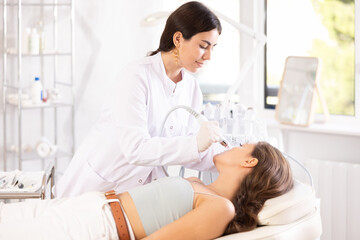 Fototapeta premium Positive young female cosmetologist performing machine-based facial procedure on client employing ion bubble pen attachment in cosmetic clinic, aimed at gentle cleanse and hydration of skin..