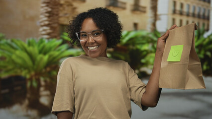 Woman wearing glasses holds paper bag with smiley note on city street; happiness generosity kindness.