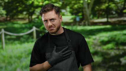 Young man with beard wearing glove and striped apron smiling outdoors in a park setting.