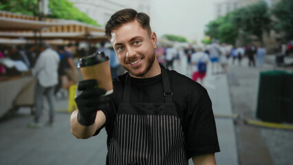 Young man with beard in apron holding takeaway coffee at outdoor market with bustling city backdrop, smiling as a waiter serving beverages in street setting.