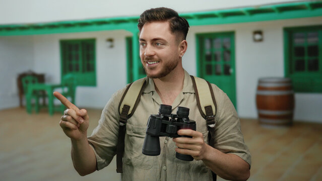 Young tourist man with a beard, holding binoculars and pointing on a city street, wears a backpack, suggesting adventure and exploration.