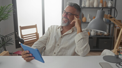 Man laughing while holding blue tablet at desk in a building office, wearing glasses and linen shirt and leaning back in chair; relaxed happiness.