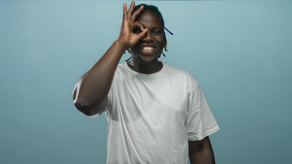 Man making ok sign with hand over eye in blue studio wearing white t shirt and open mouth...