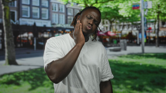 Man with hand on cheek for toothache on street near shops and trees wearing white t shirt; pain seeking help.