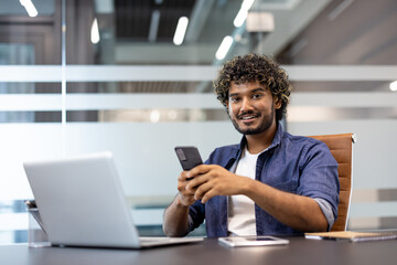 Portrait of a smiling young Indian man sitting at an office desk, holding a phone in his hand and smiling at the camera