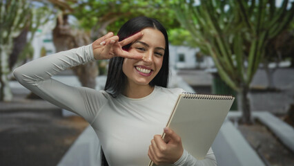Woman smiling and posing with a notebook in an outdoor park setting with trees, showcasing joy and positivity on a sunny day.