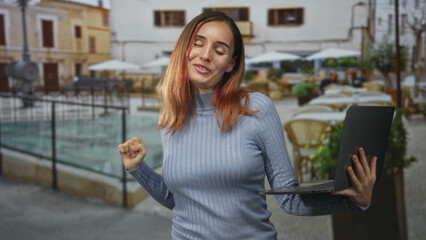 Fototapeta premium Woman holding laptop with left hand and fist pumping with right hand on street cafe, smiling and dancing while looking at screen; joy celebration.