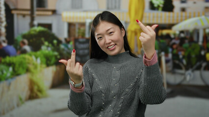 Woman gestures humorously with a playful expression outdoors in an urban setting, wearing a casual sweater, surrounded by city life and vibrant greenery, during the day.