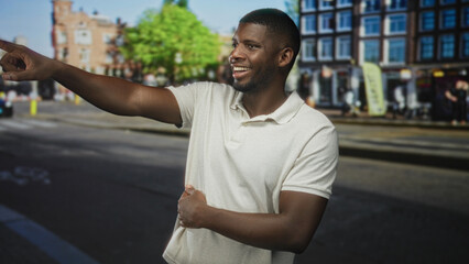 Young man points with outstretched arm while clutching stomach and showing stomachache on a busy urban street lined with buildings and cafes; concern urgency.