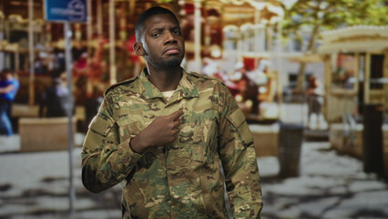 Fototapeta premium Soldier man in camouflage uniform points finger to chest in a busy street market with carousel, stalls and blurred pedestrians; pride duty.