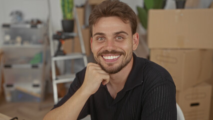 Man smiling with hand on chin in house surrounded by moving boxes and ladder and drill on shelf; moving day optimism.