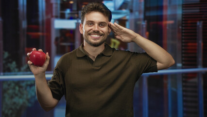 Man holding red apple salutes with right hand in hotel lobby, smiling to camera; cheerful healthy...