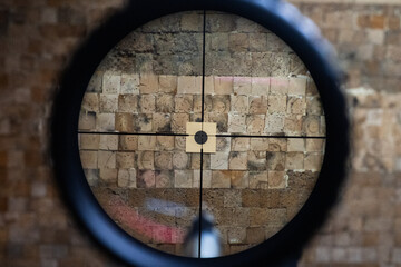 View through a rifle scope reticle aiming at a small target on a weathered wooden block wall at an indoor shooting range.