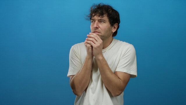 Hispanic man with worried expression clasps hands nervously against a plain blue background wearing a simple white shirt.