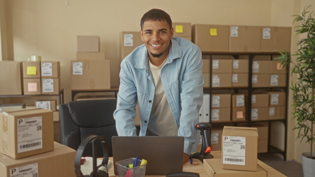 Man leaning on desk with shipping boxes and barcode scanner in building; entrepreneurial small business pride.