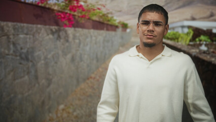 Man wearing glasses stands with hands at sides on a stone street beside a planter and wall, looking...