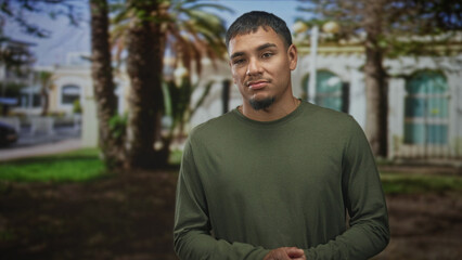 Fototapeta premium Man with hands clasped and a wry grimace, wearing an olive long sleeve shirt, standing near palm trees in front of a sunlit building; skepticism.