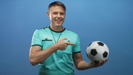 Man points finger to soccer ball with hand while holding ball and whistle visible in studio; confidence authority sport.