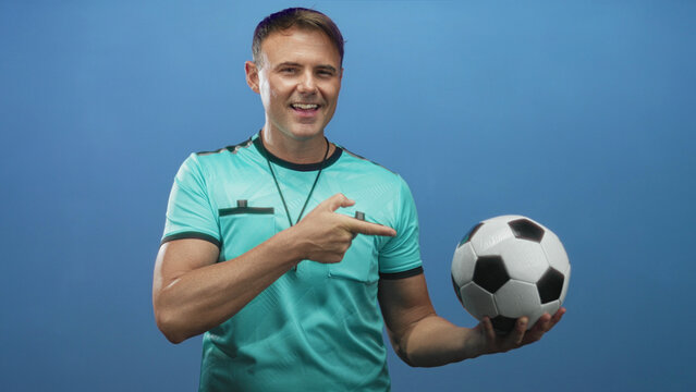 Man points finger to soccer ball with hand while holding ball and whistle visible in studio; confidence authority sport. - Powered by Adobe
