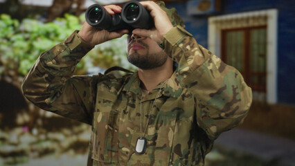 Man in camouflage uniform holding binoculars and looking upward by green foliage near building; duty vigilance.