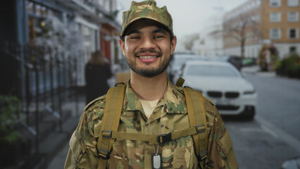 Man wearing camouflage uniform with backpack straps and dogtag smiles in urban street setting; pride duty honor.