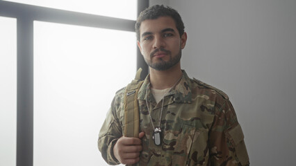 Man soldier firmly holds and adjusts backpack strap in sunlit building; commitment readiness pride duty.