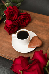 Red roses surround a coffee cup and chocolate on a wooden tray with a dark background