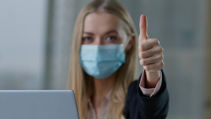 Caucasian business woman in face medical mask showing thumb up satisfied businesswoman female girl recommend safety rules coronavirus protection with laptop computer in office health care like gesture
