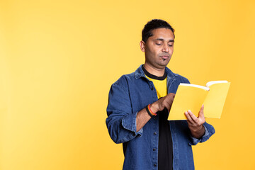 Male book lover feeling happy and pleased reading the book in studio, enjoying a fantasy plot with literature hobby. Young indian bookaholic guy browsing the novel files, book collector.