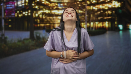Nurse woman clutching stomach with hand in scrubs on street outside illuminated building at night; amusement resilience.