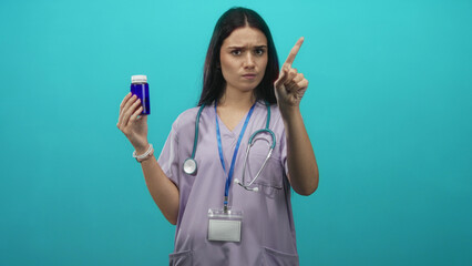 Woman nurse in lavender scrubs holding blue pill bottle, stethoscope visible and showing thumbs down gesture in studio; disapproval medication caution.