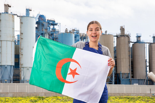 Smiling young woman in uniform posing with Algeria flag near factory