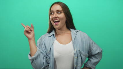Young caucasian woman pointing index finger and smiling, wearing denim shirt and white tank top in mint green studio; playful confidence.
