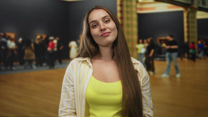 Woman smiling at camera in gallery building, close frontal pose with yellow tank top and long hair, visitors and paintings blurred behind her; quiet confidence.